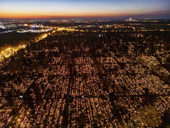 An aerial view of a cemetery illuminated with thousands of candles at dusk. The surrounding city lights and a river are visible in the background.

Widok z lotu ptaka na cmentarz oświetlony tysiącami świec o zmierzchu. Otaczające światła miasta i rzeka są widoczne w tle.