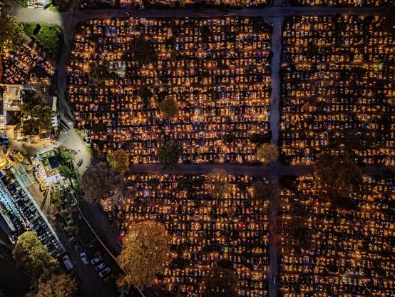 An aerial view of a cemetery at night, filled with illuminated graves that include candles and decorations. Surrounding areas feature trees and pathways, with a building visible on the left side.

Widok z lotu ptaka na cmentarz w nocy, wypełniony oświetlonymi grobami, które zawierają świece i dekoracje. W okolicy znajdują się drzewa i ścieżki, z budynkiem widocznym po lewej stronie.