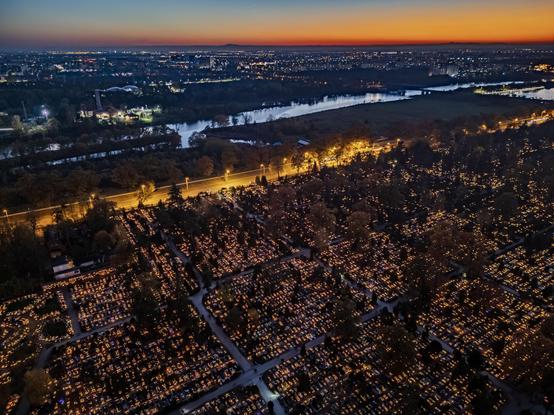 An aerial view of a cemetery illuminated by numerous candles at dusk, with a river and city lights visible in the background. The scene captures a serene atmosphere as night falls.

Widok z lotu ptaka na cmentarz oświetlony licznymi świecami o zmierzchu, z rzeką i światłami miasta widocznymi w tle. Scena oddaje spokojną atmosferę, gdy zapada noc.