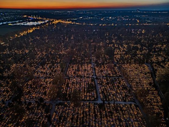 An aerial view of a cemetery illuminated by thousands of candles at dusk, with a colorful sunset and city lights in the background.

Widok z lotu ptaka na cmentarz oświetlony tysiącami świec o zmierzchu, z kolorowym zachodem słońca i światłami miasta w tle.