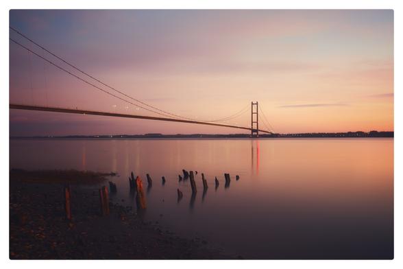 Sun sets over the Humber Bridge casting a glow all around. The remains of an old pier lead out into the river Humber from the bottom left.