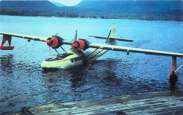 This is a big seaplane. It is parked near a dock in the wilderness. Mountains and forests are in the background behind the lake. One engine has its props turning. We might be witnessing the start of taxiing on the water.
