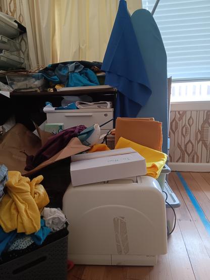 A cluttered collection of possessions in a sun room, incl but not limited to a desk, a cabinet, piles of clothing and fabric, a folded up ironing board, and a covered Janome sewing machine