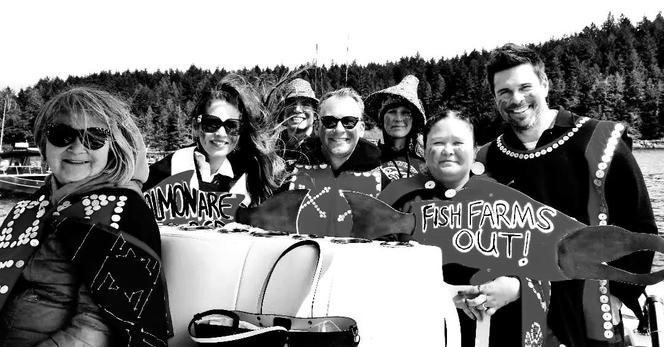 Black and white photo.
Group on boat wearing Indigenous ceremony attire & holding up fish farms out protest signs