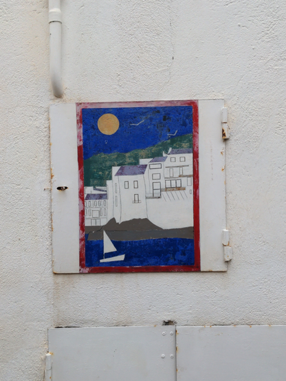 Sur une plaque technique dans un mur, est peinte une vue sur des immeubles blancs, la mer bleue au premier plan avec un voilier blanc, une colline verte et le ciel bleu au dernier plan, avec le soleil jaune.