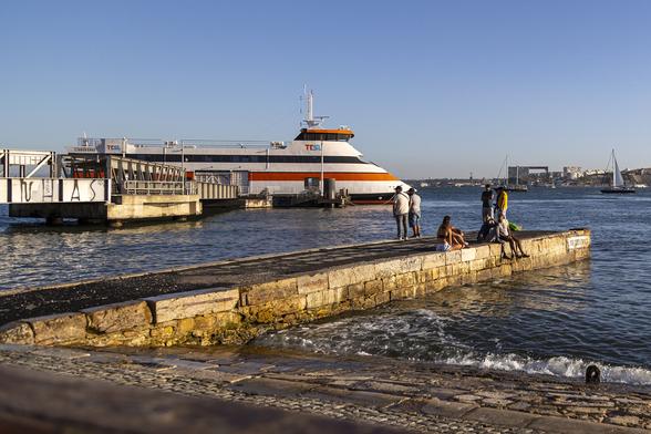 It's the early evening on a warm day with clear blue sky and light coming from the right, low down, and golden with the approach of sunset. We're standing on one shore of a wide river with a small tidal swell pushing small waves ashore up a paved stone slipway. A stone jetty stretches out and there are a handful of people at the end of it, some fishing, some simply sitting to enjoy the feel of the sun on their faces as they face to our right. Beyond, against a larger jetty with low bridge and walkway structures on it there is a passenger ferry; not very large, primarily white but with thick bands of orange and black along its hull, and with a strong slope forward at the fore from the small bridge to the waterline. It's a peaceful scene.