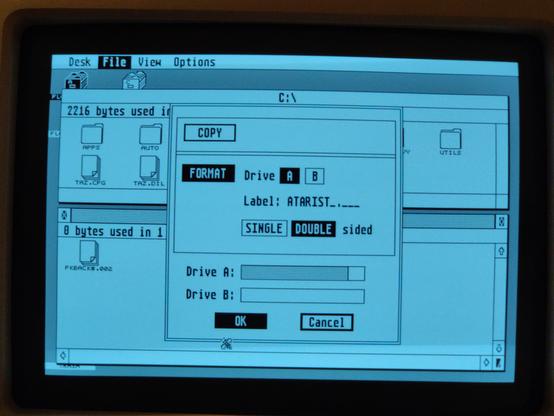 A photo of the screen of the Atari black and white high resolution monitor, showing Atari GEM's floppy format dialog. 

The dialog is divided into three panels arranged top to bottom. The first contains only a button labeled "COPY". 

The second panel has a button labeled "FORMAT" , the text "Drive" followed by two buttons with the labels "A" and "B"; a text label "Label", followed by an 8.3 format text entry field. (The text I've typed is "ATARIST") Under that are two buttons labeled "SINGLE" and "DOUBLE" followed by the text "sided".

The third panel contains two progress bars, one above the other, labeled "Drive A:" and "Drive B:" The progress bar for drive A: is at about 90%. Beneath these are two buttons, "OK" and "Cancel".