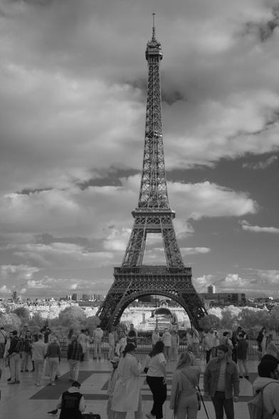 Black and white, infrared photography: Crowds at the Trocadero, the Eiffel Tower dominates the background of the frame, behind it, a sky with some clouds.
