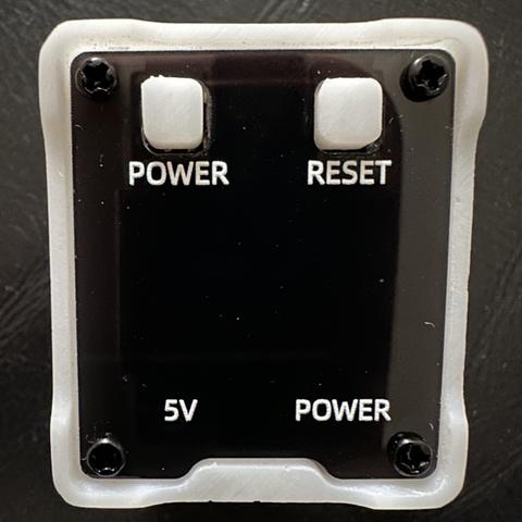 A white square surrounding a shiny black display, with 2 buttons labelled POWER and RESET and 2 labels marked 5V and POWER.