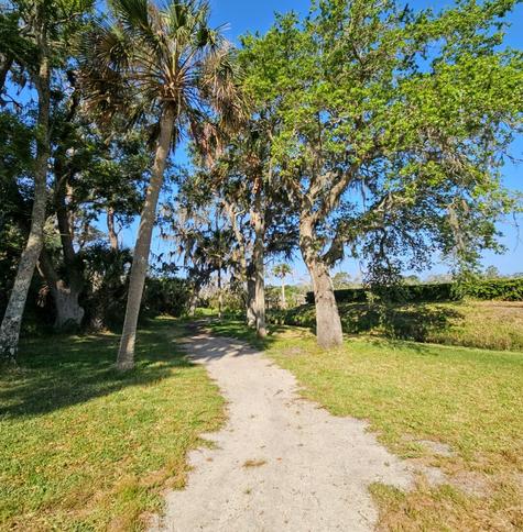 A long winding trail leads through an open area and a variety trees to an area where a large green hedge can be seen to surround something.