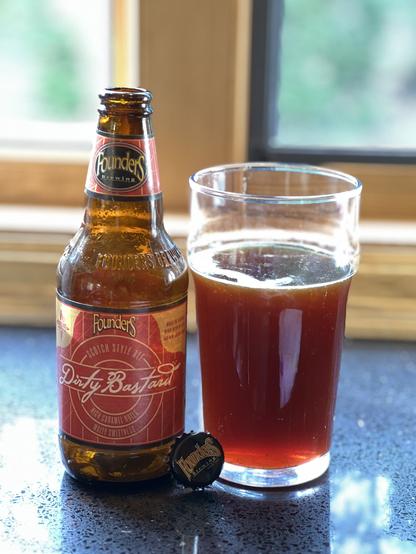 A short brown beer bottle with a red label is next to a full pint glass lit from behind by the sun, revealing a deep ruby color
