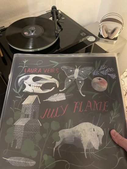 Auf einem schwarzen Plattenspieler läuft eine schwarze Schallplatte. Davor halte ich das Cover ins Bild. Es ist schwarz mit verschiedenen weißen Zeichnungen. Eine Kirche, eine Feder, ein Büffel, ein Schmetterling, ein Apfel, der Schädel eines Tiers. Außerdem sind mehrere grüne Blätter drauf. In roten Buchstaben steht Laura Veirs July Flame.