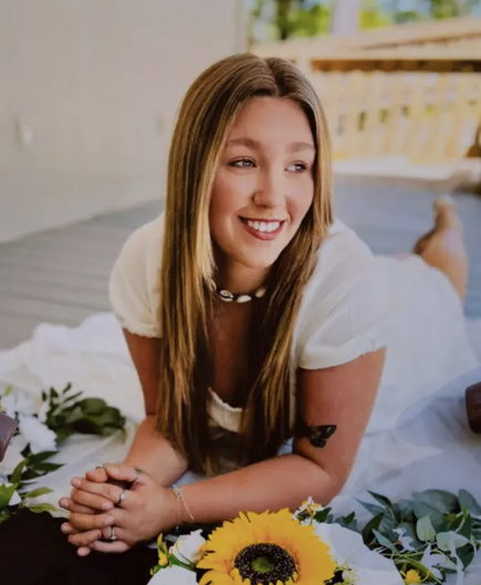 Photo of a young woman with fair skin and sandy blonde hair, posing on her elbows next to a sunflower