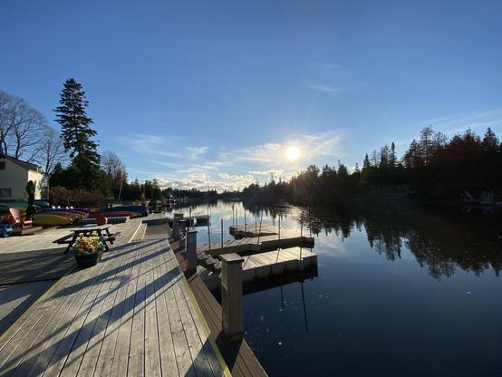 A serene riverside scene featuring a wooden dock with reflections on the water. Colorful canoes are lined up along the dock, and a picnic table sits nearby. The sun is low in the sky, casting a warm glow.