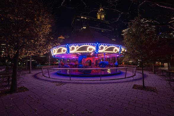 A brightly lit carousel featuring colorful animals is set against a nighttime urban backdrop, surrounded by trees with autumn leaves and modern buildings.