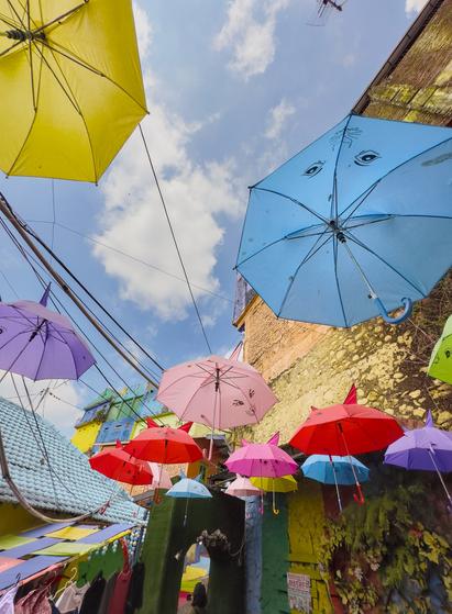 Looking up at a vibrant display of colorful umbrellas suspended between buildings. The umbrellas in yellow, red, pink, purple, and blue create a canopy against a bright blue sky with white clouds. Some umbrellas feature hand-drawn decorative eyes and patterns.