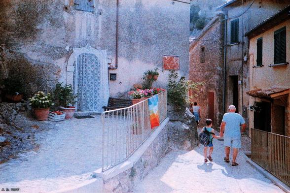 Italienische Kleinstadt in den Bergen von Lazio mit Häusern links und rechts von einer schmalen Straße, auf der ein älterer Mann und ein vielleicht 8-jähriger Junge Hand in Hand gehen. Das Haus links hat einen gepflasterten Vorhof mit Blumenkübeln und einer Regenbogenflagge am Geländer. Vor dem Eingang hängt ein Fliegengitter. Verblasste Farben und grobes Korn wegen des verwendeten Kreativfilms. Lomo LCA auf Rollei Crossbird.