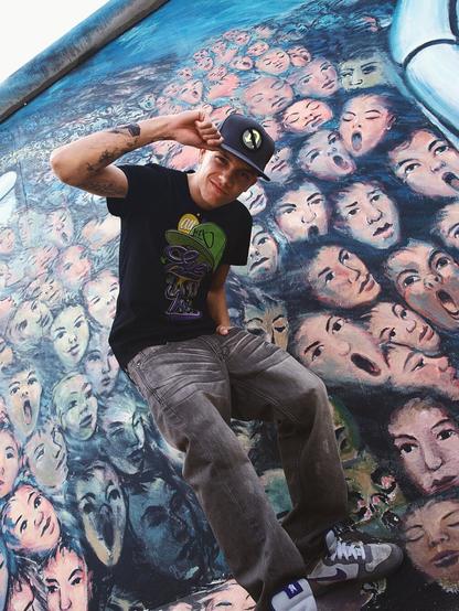 a young individual posing in front of a vibrant mural. The mural features various stylized faces and figures with dynamic expressions. The person is wearing casual attire, including a graphic t-shirt and denim pants. They are striking an energetic pose with one hand on their hip and the other raised in a wave or greeting.