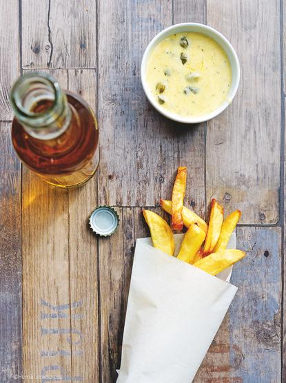 Vintage Holzuntergrund. Darauf eine Tüte aus der Backofen-Pommes-Fites herausschauen. Darüber ein Schale mit Zitronen-Kapern-Sauce. Links eine Flasche mit einem goldgelben Getränk