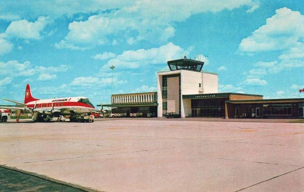 An Air Canada prop plane waits outside the terminal. There are puffy clouds against a blue sky.