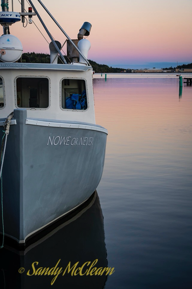 Fishing boat named “Nowe or Never” floating on flat calm water in a harbour at dusk.