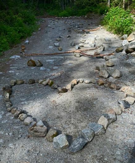 Remote logging road filled with large rocks & tree branches are spread all down the road. A large heart formed by rocks is in front. Forest surrounds the isolated road.