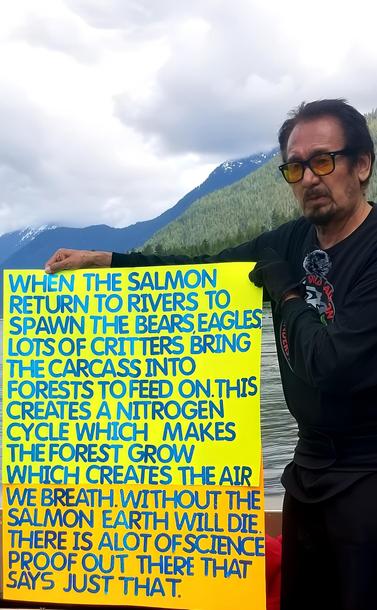 George holding up a bright neon sign that reads:

When the salmon return to rivers to spawn the bears, eagles, lots of critters bring the carcass into forests to feed on. This creates a nitrogen cycle which makes the forest grow. Which creates the air we breathe. Without the salmon Earth will die. There is a lot of science proof out there that says just that.