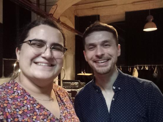 selfie of myself (left) and Adam Neely (right), both smiling. I am wearing a floral dress and big golden earrings, and a Star of David necklace. Adam a dark-blue v-neck shirt.