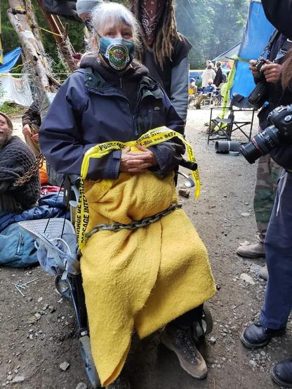Jane, in her walker with yellow police tape & chained to a hardblock tripod at a logging road blockade. Other activists are on ground, chained to same tripod block. Journalists with cameras are on the right. Activist support teams are in background.