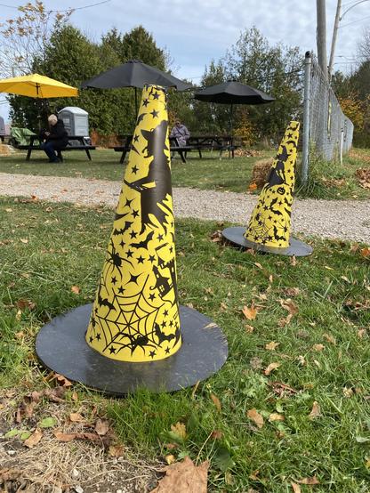 Two yellow traffic cones with Halloween-themed designs, featuring witches, spiders, and bats, are placed on a grassy area. In the background, there are picnic tables with black umbrellas.