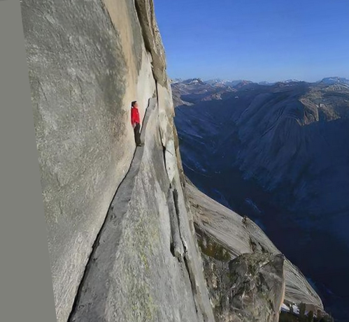 Color image of a hiker following a ridge path on a rock face as it narrows. Rock face is left in the image, tilted back from upright perhaps 20 degrees, ledge is narrow and ascending, image beyond is blue sky and a mountain range.