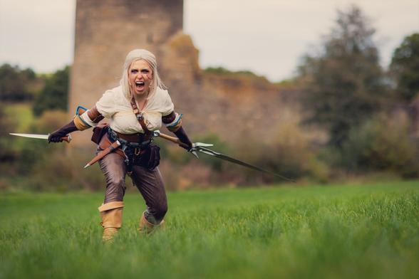 A cosplayer dressed as Ciri from The Witcher series charges forward with two swords in hand, wearing her iconic white tunic and leather armor, exuding power and determination in a battle-ready pose.