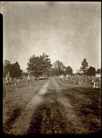 Black and white but slightly sepia toned image of a straight gravel path through a historic cemetery. A large tree is in the distance.
