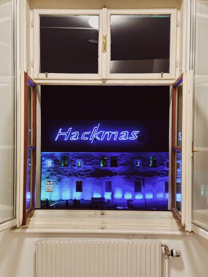 Das Bild zeigt ein Fenster, wo beide Fenstertüre geöffnet sind. Es zeigt einen Blick hinaus in die dunkle Nacht zum gegenüberliegenden Schlosswand/-dach. Auf dem Dach ist mit Laser angeleuchtet "Hackmas" im grellen blau/lila-Tönen. Darunter ist die Schlosswand in selbigen Farben angeleuchtet.