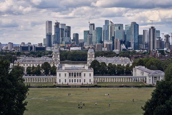The image depicts Greenwich Park in London, with the historic Queen's House and the Old Royal Naval College in the foreground. In the background, you can see the modern skyscrapers of Canary Wharf. The scene combines lush green parkland with historical and contemporary architecture.