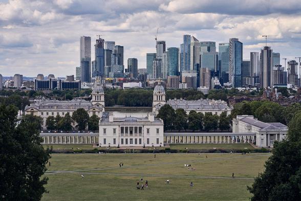 The image depicts Greenwich Park in London, with the historic Queen's House and the Old Royal Naval College in the foreground. In the background, you can see the modern skyscrapers of Canary Wharf. The scene combines lush green parkland with historical and contemporary architecture.