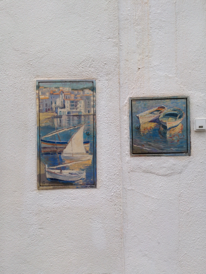 Sur deux plaques adjacentes dans un mur blanc, deux peintures, sans doute reproductions impressionnistes : celle de gauche représente une plage où se reposent trois petits bateaux, devant des immeubles blancs dans le fond, signé "CREA" ; le deuxième deux barques dans la mer devant la plage.
