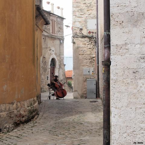Farbfoto, aufgenommen in einer engen Gasse der Altstadt von Narni, Umbrien. Zwischen den Häusern links und rechts spielt auf dem Straßenpflaster ein Cellist sein Instrument. Digitalfoto, Olympus E-M5, Zuiko 14-42 mm.