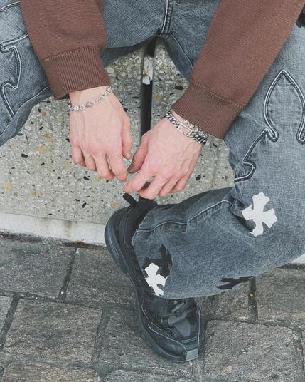 Bang Chan showing his regular round flower cross bracelet, plus a new bracelet with the same symbol interspersed with others. And wearing jeans that have the flared cross symbol.