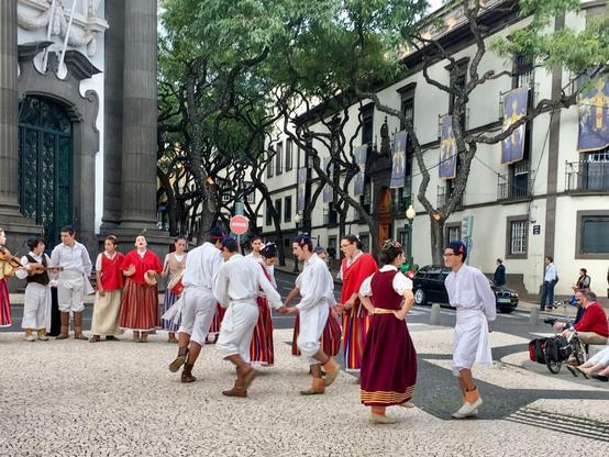 Das Bild zeigt eine Gruppe junger Menschen bekleidet in madeirensischen Trachten, die in Funchals Fussgängerzone tanzen. Die Männer in weiß, mit schwarzer Kappe, die Frauen in rotbraunen Kleidern. Begleitet werden sie von einer Gruppe dabei stehender Musiker und Sänger.