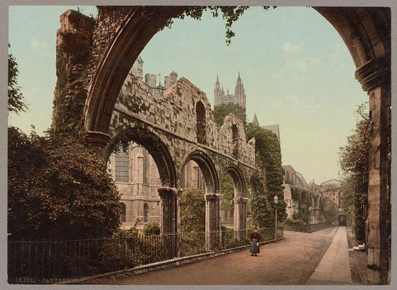 The image depicts the ruins of St. Augustine's Abbey in Canterbury, England. The structure features an imposing archway with large stone pillars and intricate detailing on its surface. Vines cover parts of the wall, adding a sense of age and decay to the scene.

In the background stands another historic building, possibly part of a cathedral or castle, with spires visible against the sky, indicating that this location is rich in cultural heritage. The setting appears serene and quiet, as we see one solitary figure walking along a paved path lined by metal railings on either side.
The overall color scheme suggests an early 20th-century photograph, likely produced using photochrom printing techniques common during that era for creating images from printed photographs onto paper or card stock.