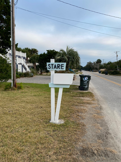 Color image of a rural streetside mailbox on a South Carolina coastal community. The image shows a white mailbox on a white and pale blue wood post, with the single word, "STARE", on a placard above the mailbox. Cloud-mottled blue sky above, a rural street on the right side of the image, yards, cars, and trees.