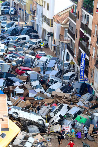 A street in Valencia, Spain, blocked in 2024 by the debris and detritus left by flash flooding likely exacerbated by Climate Change. The street is piled high with wrecked vehicles tossed willy-nilly like olives, capers and tomatoes in a Spanish salad.