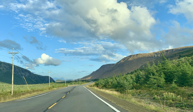 A two lane road with mountains on either side. The mountain on the right is made of orangish rock. There is some grass and low brush in the foreground on either side of the road.