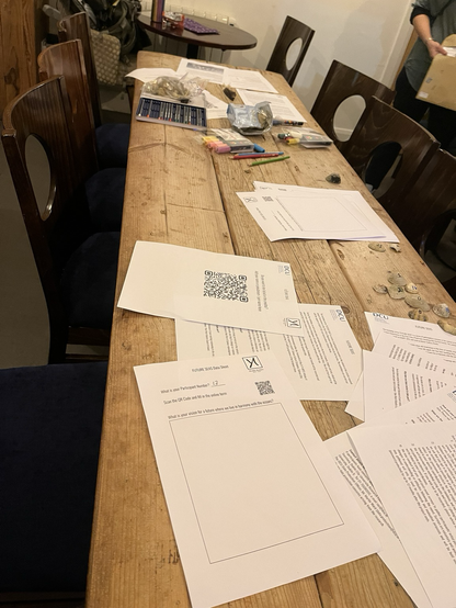 A long wooden table has papers and materials for the workshop strewn over its surface. In the right of the table are sea shells with numbers painted on them.