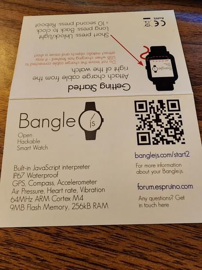 Instruction card for the Bangle.js smartwatch on a wooden surface. The card provides quick-start instructions for charging and basic operations of the smartwatch. Top half displays a diagram showing how to attach the charge cable to the right side of the watch. Instructions include pressing options: 'Short Press: Unlock/Light,' 'Long Press: Back to Clock,' and '10 second press: Reboot.' Bottom half lists features of the smartwatch: 'Open, Hackable Smart Watch,' with a built-in JavaScript interpreter, waterproof rating (IP67), GPS, compass, accelerometer, air pressure, heart rate, vibration sensor, 64MHz ARM Cortex M4 processor, 9MB flash memory, and 256kB RAM. QR code links to the start guide at 'banglejs.com/start2' and community forum at 'forum.espruino.com' for further assistance.