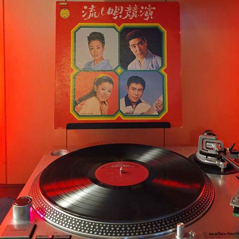 A vinyl record sits on a turntable. Behind the turntable, a vinyl album outer sleeve is displayed. The front cover shows 4 different Japanese singers (Hibari Misora, Hideo Murata, Harumi Miyako, and Saburo Kitajima)