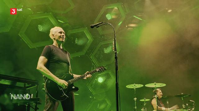 Ausschnitt aus einer 3sat-Reportage: Zu sehen ist eine grün-gelbliche Bühne der unwiderlegbaren besten Band der Welt, die ärzte aus Berlin... aus Berlin! Links im Vordergrund spielt Farin Urlaub Gitarre. Er hat ein – natürlich schwarzes – Shirt, spielt auf einer schwarzen Gitarre und lächelt zum Publikum. Rechts im Hintergrund ist der Schlagzeuger Bela B zu sehen.