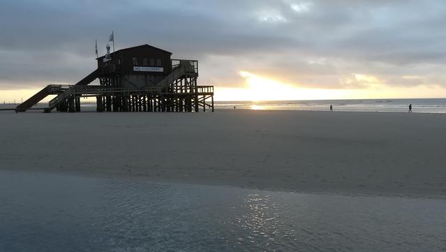 Nordseestrand bei untergehender Sonne am wolkigen Himmel. Im Hintergrund ist ein sogenannter Pfahlbau zu sehen. Diese sind typisch für Sankt Peter Ording.
