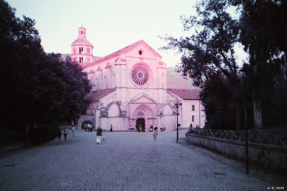 Farbfoto der Abteikirche des Klosters Fossanova in der Provinz Latina, Lazio, Italien, erbaut um 1208 im Stil der frühen Gotik. Zu der majestätischen Kirche führt eine breite gepflasterte Straße, die von Bäumen und rechts einer niedrigen Mauer gesäumt ist. Verfremdete Farben wegen des verwendeten Purple-Films.